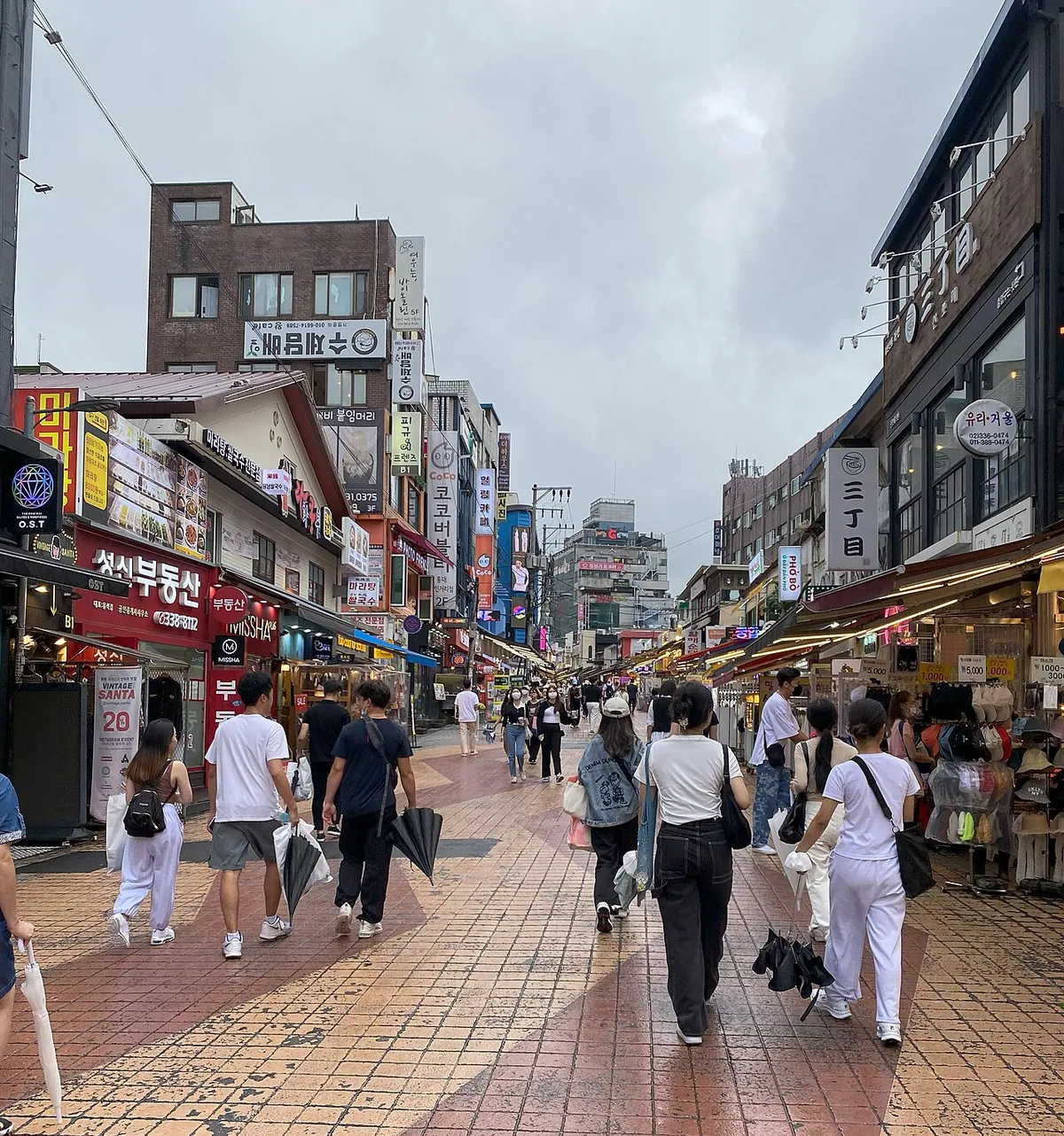People walking along a bustling street lined with shops in Hongdae, Seoul.